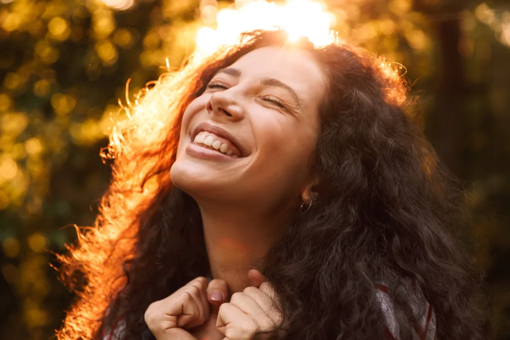 Portrait closeup of happy curly woman 18-20 lsmiling with closed eyes and expressing delight while walking through park in sunny day with trees background