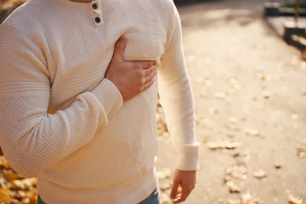 Moung man stands on a park alley and holds his hand to his sternum, he is in pain